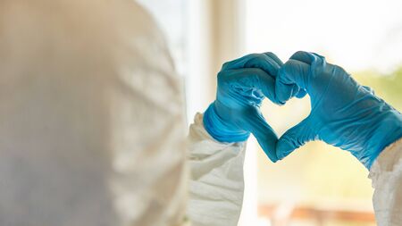 Clinical staff in protective clothing holds heart with his hands in front of window as corona virus...
