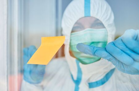 Yellow sticky note slip on door or window of clinic with doctor in protective clothing during coronavirus epidemicの写真素材