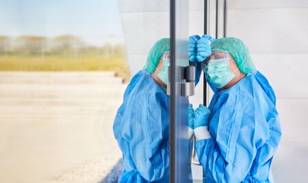 Exhausted doctor in protective clothing leans on door because of stress and exhaustion from coronavirus and Covid-19 pandemicの写真素材