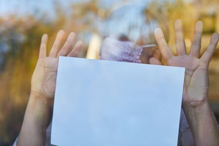Lonely elderly woman with a face mask holds notes on window in coronavirus quarantine because of...