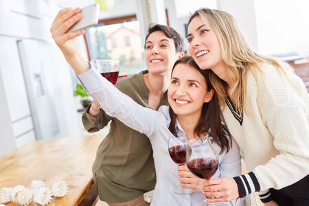 Three happy girlfriends with red wine take selfie together in the kitchen of a shared apartment