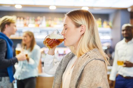 Young woman drinking with pleasure a glass of beer in the pub with friends in the backgroundの写真素材
