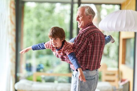Grandfather makes happy grandson fly at home in the living roomの写真素材