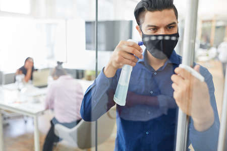 Businessman with mask because of Covid-19 disinfecting glass with virucidal cleaning agent in the officeの写真素材