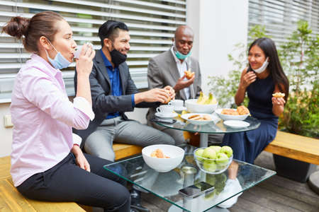 Young business people during a break eating together at the table with mouth-nose protection on the...