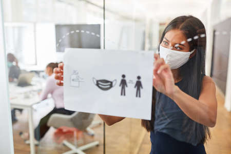 Business woman with everyday mask fastens notice board with rules of conduct against infection with Covid-19の写真素材