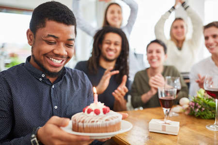 Happy african man with cake and candle at birthday with many friends at homeの写真素材