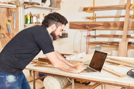 Young craftsman in the workshop at the laptop. Computer writes an emailの写真素材