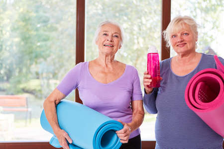 Two vital senior women with yoga mat in yoga class at the fitness centerの写真素材