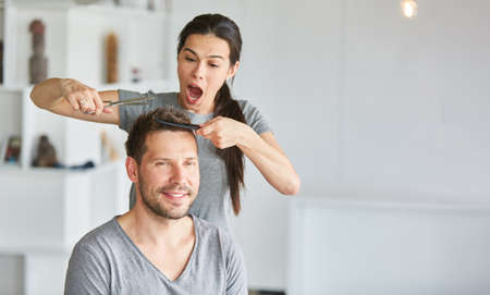 Young woman has fun while cutting her partner's hair with scissors and combの写真素材