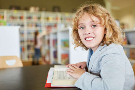 Happy blond boy is reading in a book in the library of elementary schoolの写真素材