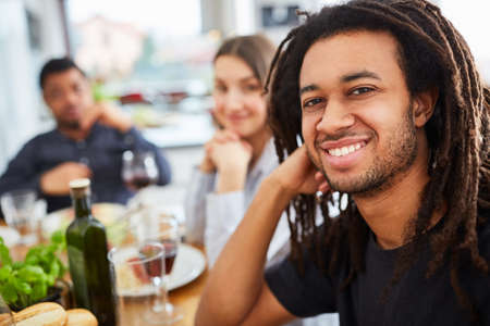 African man with dreadlocks sits with friends at the dining table having a mealの写真素材