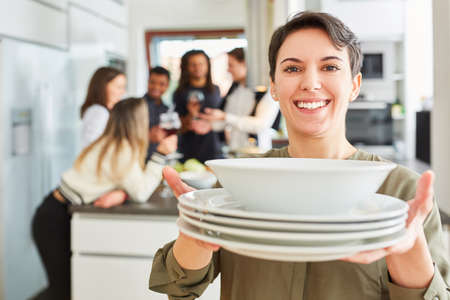 Young woman with dishes at table setting in front of student friends in shared kitchenの写真素材