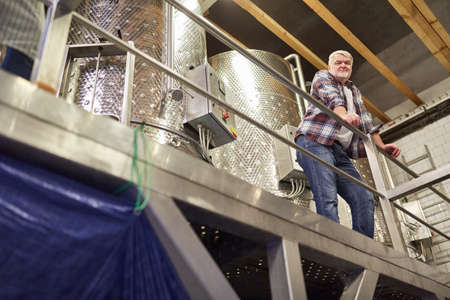 Older winemaker as master winemaker monitors vinification in front of a fermentation tank
