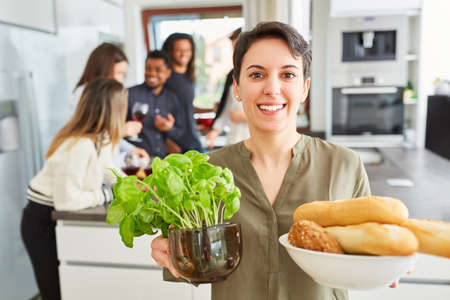 Woman with pot of basil and bowl of baguette in a shared kitchen with friends in the backgroundの写真素材