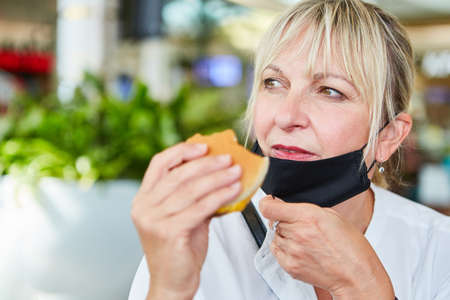 Woman as a guest with a face mask on her chin while eating a burger in a fast food restaurant