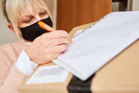 Woman with face mask in front of apartment door gives delivery service a signature for package