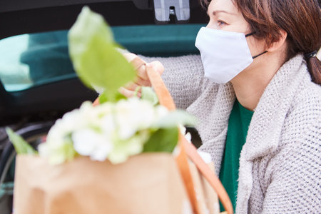 Woman with surgical mask carries bag with flowers in hands out of car trunkの写真素材