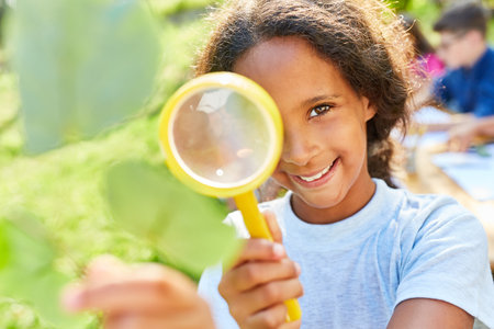 Happy girl in summer school at summer camp looks through magnifying glass at a sheetの写真素材