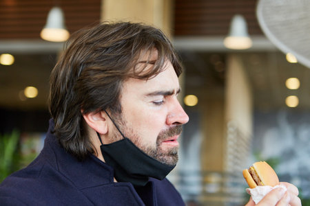 Man with a mask on his chin because of Covid-19 while eating a burger in the snack restaurantの写真素材