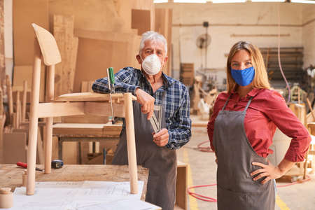 Senior craftsman and woman as an apprentice with face mask in the cabinet makerの写真素材