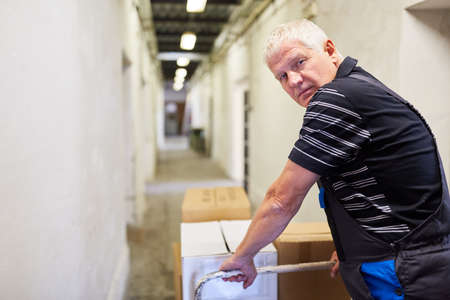 Elderly worker pushes packages on a push cart to the incoming goods department of the factoryの写真素材