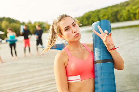 Sporty and casual young woman with yoga mat after fitness class on the lakeside