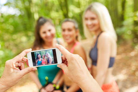 Hands holding smartphone while taking a picture of three women doing Nordic walkingの写真素材