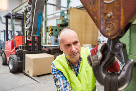 Worker in a warehouse attaches load to crane hook with forklift truck in background