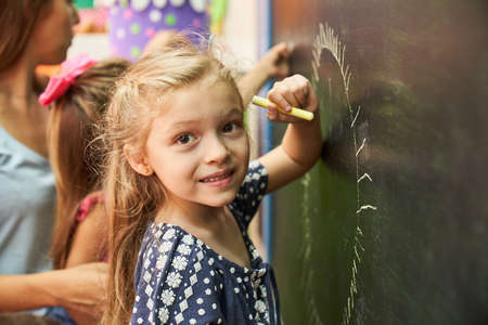 Little girl in preschool or kindergarten while painting with chalk on the blackboardの写真素材