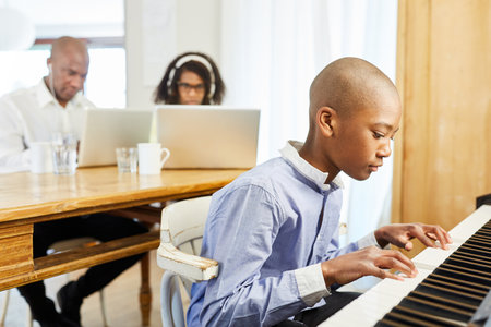 Child is practicing on the piano in the living room as a home office with parents at the computer in the backgroundの写真素材