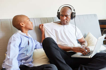 Child disturbs annoyed father during a video conference on the laptop computer on the sofa in the home officeの写真素材