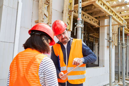 Foreman and architect with tablet computer in front of the shell of a building site for building a...