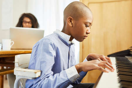 African boy learning to play piano in living room with mother on laptop pc in backgroundの写真素材