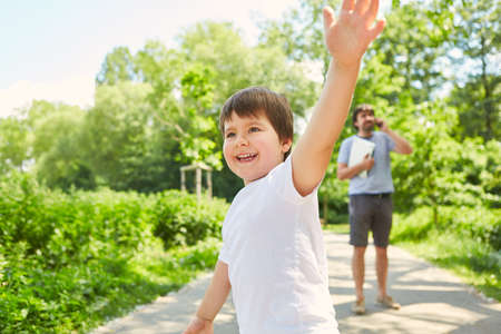 Father talking on mobile phone in park in summer with son playing in foregroundの写真素材