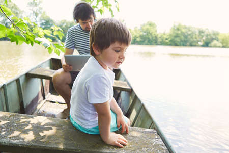 Boy in boat on a lake on summer vacation with father on laptop in backgroundの写真素材