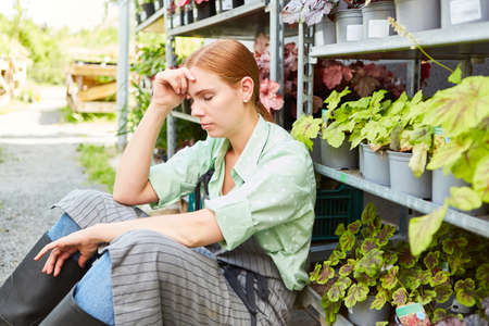 Young woman as a gardener sits exhausted in the nursery with flowers and plants