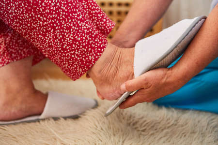 Nursing service woman or geriatric nurse helps senior citizen put on slippers at homeの写真素材