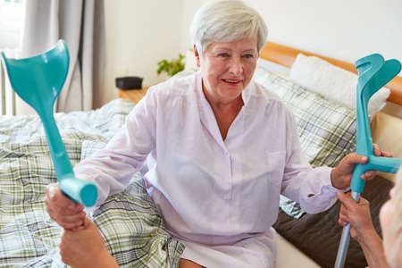 Smiling senior man in bed as a patient with crutches being looked after by geriatric nurses in the retirement homeの写真素材
