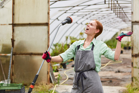 Young woman as a gardener trainee sings exuberantly into a hose in the nurseryの写真素材
