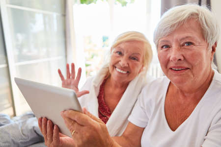 Happy senior citizen waves at the webcam during video chat with the support of a caregiver