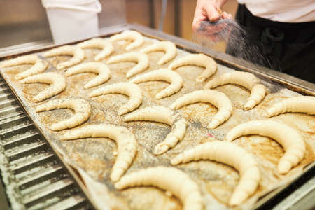 Hands of baker sprinkle croissants on the baking tray with poppy seeds in bakery or bakery factory