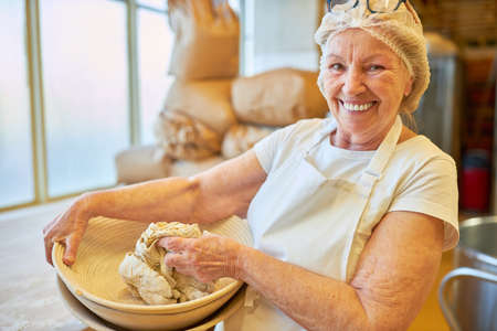 Happy elderly woman baker kneading dough in a bowl in preparation for bakingの写真素材