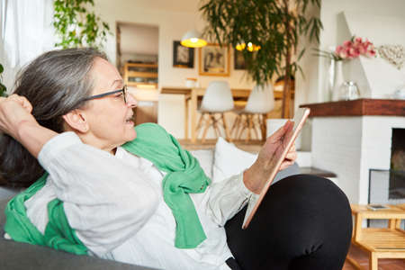 Old woman on the sofa at home or in retirement home video chatting on the tablet computerの写真素材