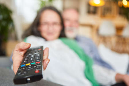 Elderly woman holds the TV remote control in her hand as a symbol for television consumption and...