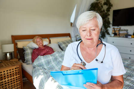 Female doctor looks at patient files in nursing home during home visit with senior citizen in the backgroundの写真素材