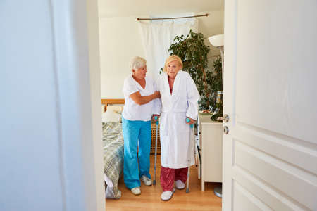 Caregiver helps senior woman in a bathrobe with crutches while walking in the rehab clinic or at...