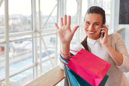 Young woman with many shopping bags waving and talking on the cellphone after shopping