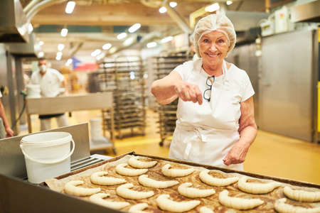 Elderly woman as an experienced baker sprinkles croissants with poppy seeds in a bakery or large-scale bakeryの写真素材