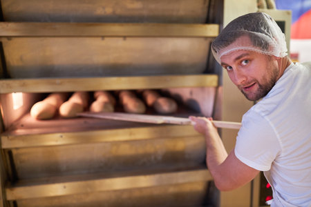 Baker in the bakery or bakery with a bread pusher on the oven while baking bread